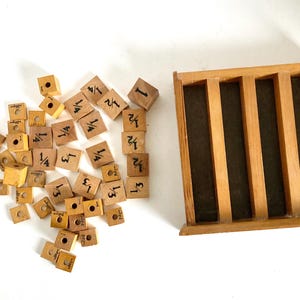 May include: A collection of small, wooden blocks with fractions and numbers, alongside a wooden tray with vertical dividers. The blocks are light brown with black markings. The tray is also light brown.