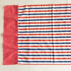 May include: A rectangular pillowcase with a red border on the left side and a striped pattern. The stripes alternate between red, white, and blue. The pillowcase is made of fabric and is likely for a bed.