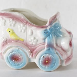 May include: A vintage ceramic baby carriage planter in pastel colors. The carriage is white with pink and blue accents, featuring flower and bow details. A yellow bird is perched on the side, and the wheels have blue and pink floral designs.