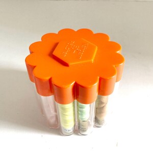 May include: An orange, flower-shaped container with clear tubes holding spools of thread. The container has a hexagonal emblem on top with text. The thread spools are various colors, including yellow, green, and brown.