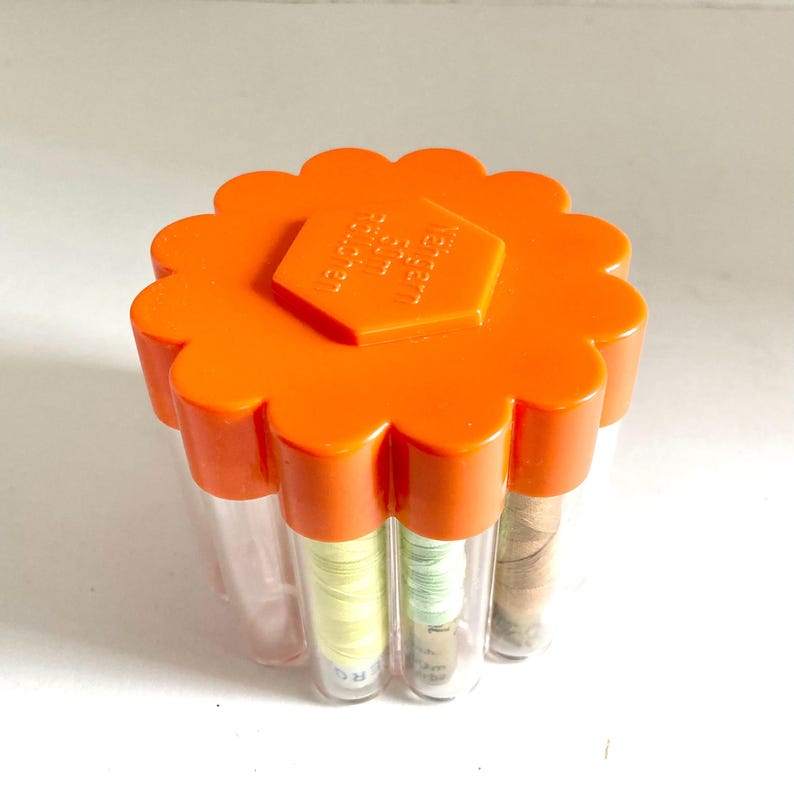May include: An orange, flower-shaped container with clear tubes holding spools of thread. The container has a hexagonal emblem on top with text. The thread spools are various colors, including yellow, green, and brown.