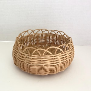 May include: A round, woven wicker basket with a scalloped edge. The basket is light brown and has a simple, natural design. It is suitable for storage or display.