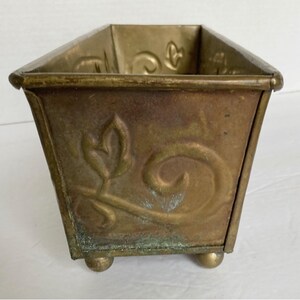 May include: A gold metal planter with a decorative floral design and four feet. The planter is rectangular and has a slightly distressed finish.