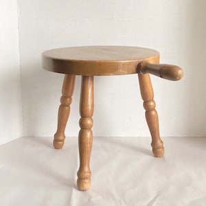 May include: A wooden stool with a round seat and three turned legs. The stool has a single handle on the side.