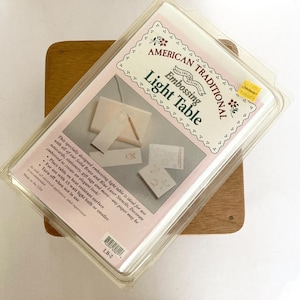 May include: An American Traditional brand embossing light table in clear plastic packaging. The product label features the text 'American Traditional Embossing Light Table' and includes an illustration of the device with stencils and a stylus.