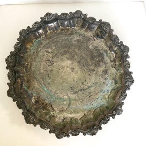 May include: A round silver-toned metal serving tray featuring a scalloped edge with ornate embossed detailing. The center of the tray displays a tarnished surface with visible oxidation, green patina, and faint etched swirling patterns.