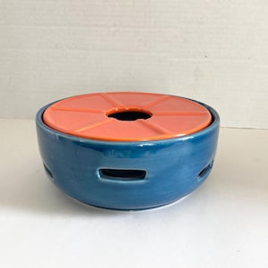 May include: A round, ceramic tealight warmer with a coral-colored lid. The lid has a circular opening in the center and is divided into segments. The base has a series of small, horizontal openings. The warmer is designed to hold a tealight candle.