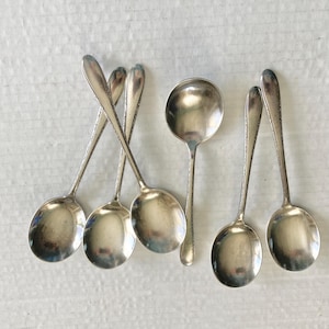 May include: A collection of silver spoons with oval bowls and long, textured handles. The spoons are arranged on a white surface, with some overlapping. The spoons are likely for serving or eating.