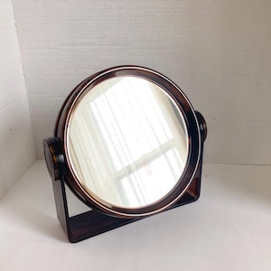 May include: A vintage, round vanity mirror with a dark brown, tortoiseshell-style frame. The mirror is supported by a matching dark brown stand. The mirror reflects the surrounding environment.