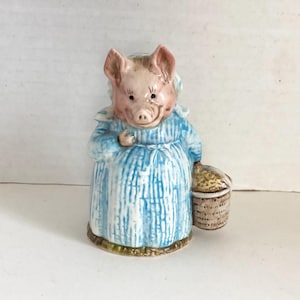 May include: A ceramic figurine of a pig wearing a blue dress with white vertical stripes and a white bonnet. The pig is holding a woven basket. The figurine has pink ears and snout, and is standing on a green base.