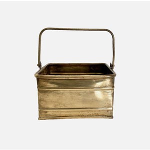 May include: A rectangular, antique brass bucket with a curved handle. The bucket has a slightly aged appearance, with visible wear and tear. The handle is attached to the top edges of the bucket. The overall design is simple and functional.
