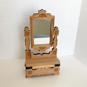May include: A small, wooden vanity mirror with a rectangular mirror and decorative carvings. The mirror is framed by a light brown wood with starburst and geometric designs. The base has a small shelf and drawer.