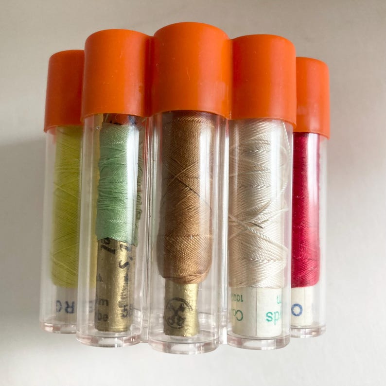 May include: A collection of six spools of thread in clear plastic tubes with orange caps. The threads are various colors, including green, brown, cream, and red. The spools are arranged together, showcasing the variety of colors.