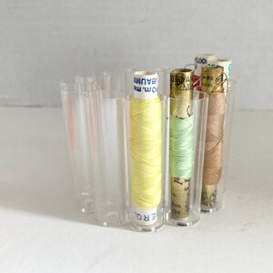 May include: A clear acrylic thread organizer holding several spools of thread in various colors. The spools include yellow, light green, and brown thread. The organizer has a curved design with individual compartments for each spool.
