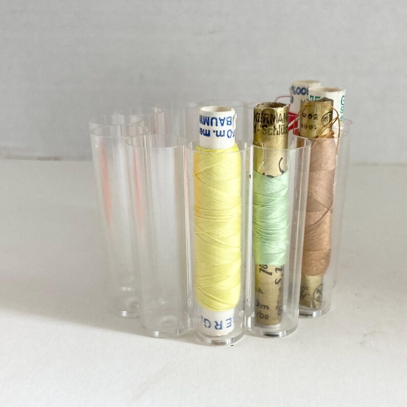 May include: A clear acrylic thread organizer holding several spools of thread in various colors. The spools include yellow, light green, and brown thread. The organizer has a curved design with individual compartments for each spool.