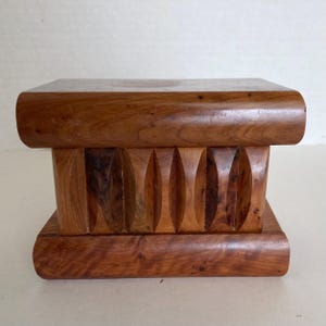 May include: A rectangular wooden display stand featuring a smooth top and base with rounded edges. The central section consists of a series of vertical, carved, almond-shaped indentations that create a textured, rhythmic pattern across the front.