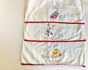May include: A white fabric item with three panels, each featuring embroidered figures and text. The panels are separated by red stripes. The top panel reads "THEY WED," the middle "HE PROPOSED," and the bottom "THEY MEET."