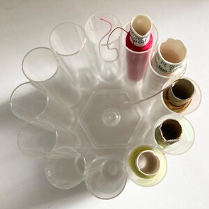 May include: A clear, circular thread organizer with cylindrical compartments. Some compartments hold spools of thread in various colors, including red and yellow. The organizer is made of transparent plastic.