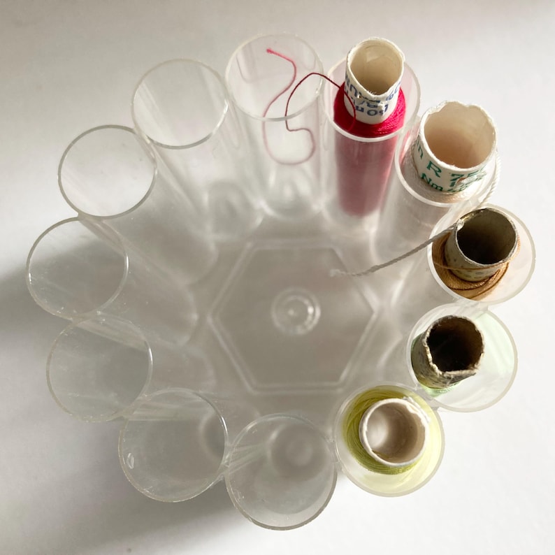 May include: A clear, circular thread organizer with cylindrical compartments. Some compartments hold spools of thread in various colors, including red and yellow. The organizer is made of transparent plastic.