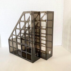 May include: A pair of translucent dark brown plastic magazine holders featuring a grid-like cutout pattern on the sides. Each holder has an open top and an angled front edge, designed for organizing documents, files, or magazines on a flat surface.