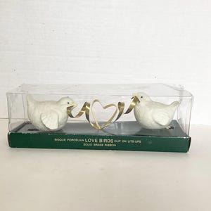 May include: Two white bisque porcelain birds in a clear plastic box with a dark green base. The birds are connected by a gold-colored solid brass ribbon shaped into a heart. The box is labeled Bisque Porcelain Love Birds Clip on Lite-Ups.