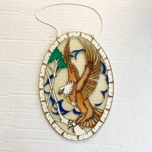 May include: Oval-shaped stained glass art piece featuring a brown eagle in flight. The eagle has white and yellow accents, set against a backdrop of a tree and blue accents. The artwork is framed with a white and brown border and includes a hanging string.
