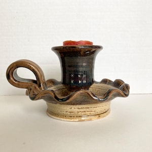 May include: A ceramic candle holder with a dark brown glazed top and a light beige base. The piece features a ruffled edge, a side handle, and a central cylindrical cup containing a small red wax candle. The surface has a glossy finish.
