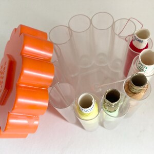 May include: A clear plastic sewing thread organizer with an orange lid. The organizer has multiple cylindrical compartments holding spools of thread in various colors, including red, yellow, and white. The lid has text.