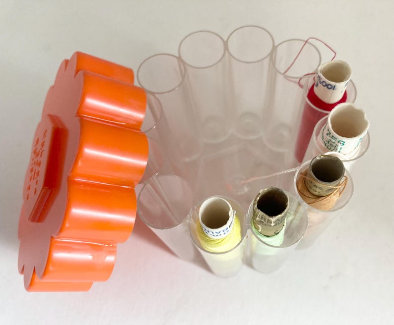 May include: A clear plastic sewing thread organizer with an orange lid. The organizer has multiple cylindrical compartments holding spools of thread in various colors, including red, yellow, and white. The lid has text.