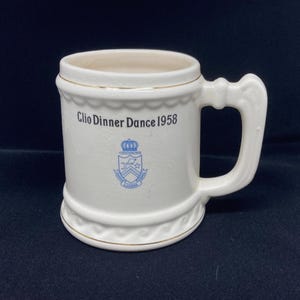 May include: White ceramic mug with a large handle, featuring the text "Clio Dinner Dance 1958" and a blue crest. The mug has gold trim around the top and bottom, and a decorative scalloped design.