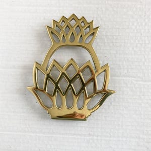 May include: A gold-colored metal trivet with a floral design. The trivet features a stylized flower shape with intricate cut-out patterns, including diamond and teardrop shapes. The design is symmetrical and has a handle at the top.