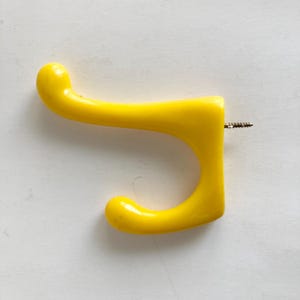 May include: A bright yellow wall hook with a curved design. The hook is made of a glossy material and includes a screw for mounting. The hook is designed for hanging items.