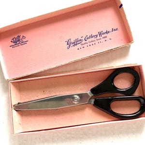 May include: Vintage Griffon Cutlery Works scissors in a pink box. The scissors have black handles and a silver blade. The box is pink with the Griffon Cutlery Works logo and text printed on the inside of the lid.