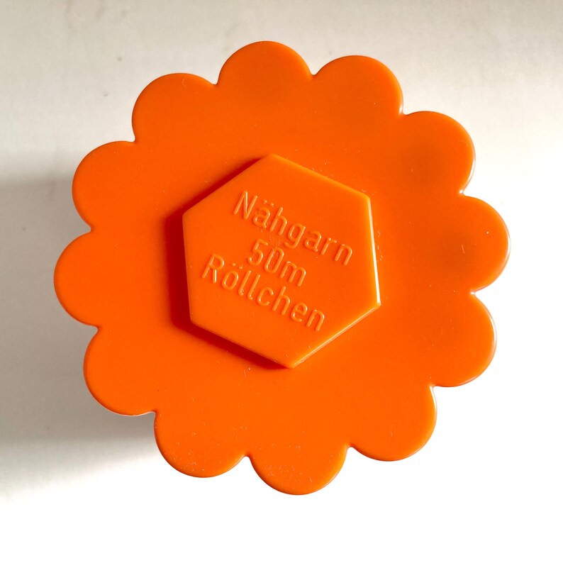 May include: Bright orange spool of thread with a flower-shaped design. The center features a hexagonal label with the text "Nahgarn 50m R&ouml;llchen". This sewing supply is ideal for various crafting projects.