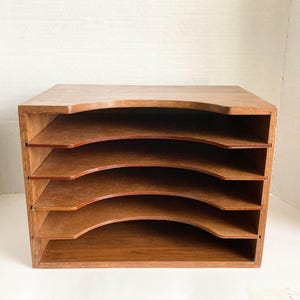 May include: A wooden desk organizer with five tiered compartments. The organizer is made of light brown wood and has a curved design on the front of each shelf. The top of the organizer is flat.
