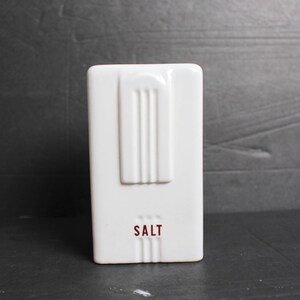 May include: A white rectangular salt shaker with a vertical ribbed design and the word "SALT" in red lettering. The salt shaker has a retro, art deco style.