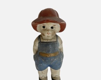 Pode incluir: Figura de cerâmica vintage de uma criança usando macacão azul, chapéu marrom e sapatos pretos. A figura tem um rosto pintado com uma expressão ligeiramente triste. A estética geral sugere um item colecionável ou decorativo.