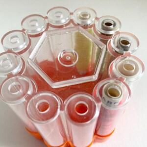 May include: A clear plastic thread spool holder with a hexagonal center and cylindrical compartments. The holder contains several spools of thread, some with red thread and others with white thread. The base is orange.