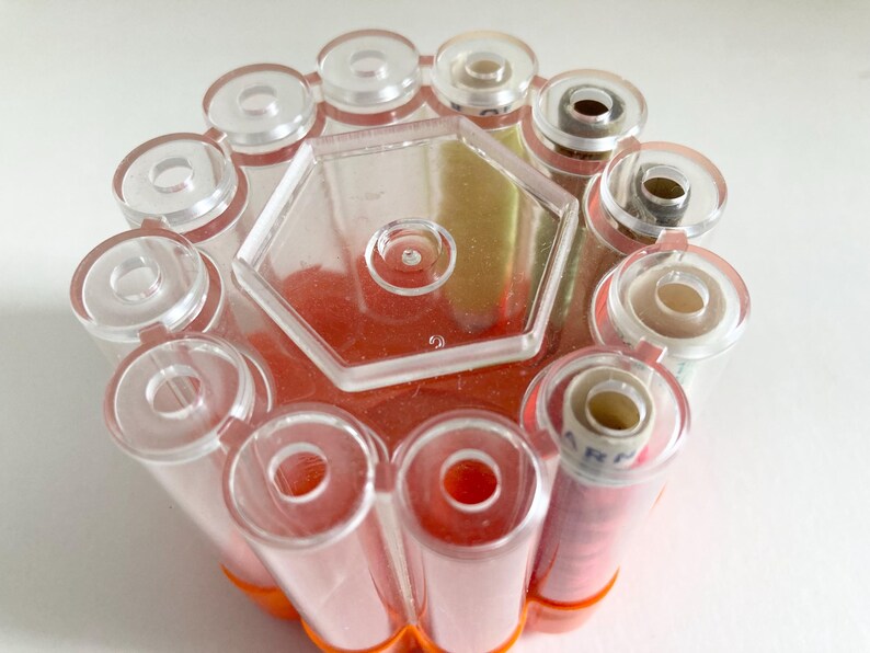 May include: A clear plastic thread spool holder with a hexagonal center and cylindrical compartments. The holder contains several spools of thread, some with red thread and others with white thread. The base is orange.