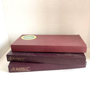 May include: Three vintage Scrabble crossword game boxes. The top box is a deep red, with a circular sticker. The two lower boxes are a darker burgundy, with the word "SCRABBLE" in gold lettering. The boxes are stacked on top of each other.