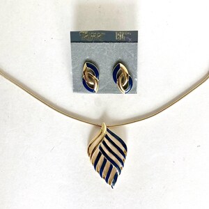 May include: A gold-tone necklace with a leaf-shaped pendant featuring navy blue enamel stripes, paired with matching stud earrings on a Trifari display card labeled Surgical Steel Posts. The jewelry set displays a consistent metallic finish.