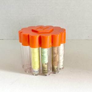 May include: A vintage orange plastic thread organizer with a flower-shaped lid. The organizer holds several clear tubes filled with colorful spools of thread. The thread colors include yellow, green, brown, and white.