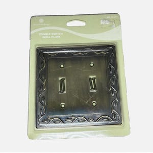 May include: A double switch wall plate in a bronze finish, featuring a decorative leaf design around the edges. The plate is packaged in a clear plastic blister pack with the words "DOUBLE SWITCH WALL PLATE" printed on the packaging.