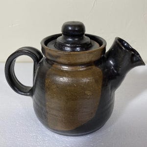 May include: A handcrafted ceramic teapot with a dark brown and black glaze. The teapot features a rounded body, a curved handle, and a spout. The lid has a small knob for easy removal. The teapot is suitable for brewing tea.
