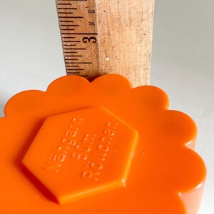 May include: Close-up of an orange, scalloped-edge sewing tool with a hexagonal center. A wooden ruler is placed on top, showing the 3-inch mark. The tool has the text "N&auml;hern F&uuml;r Rollen" on the center.