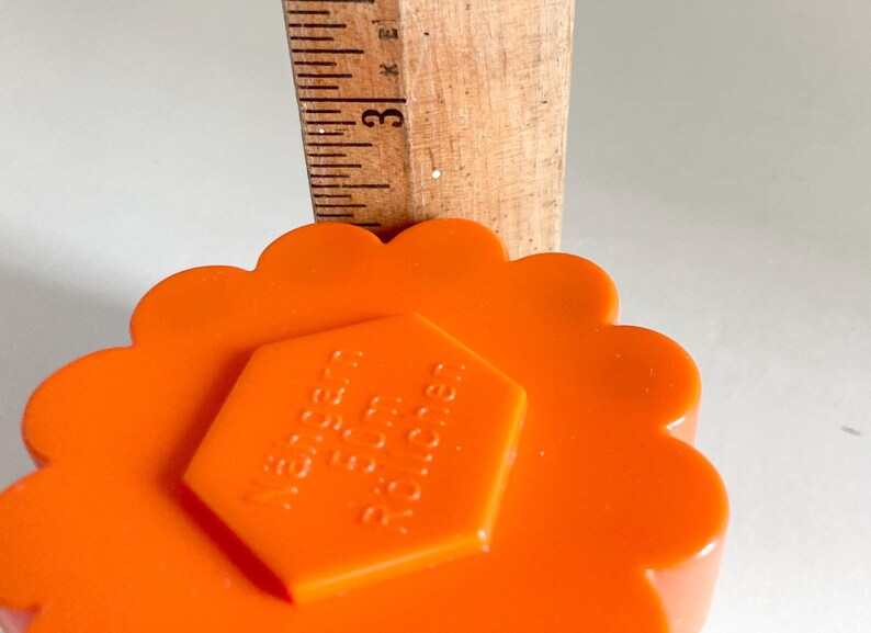 May include: Close-up of an orange, scalloped-edge sewing tool with a hexagonal center. A wooden ruler is placed on top, showing the 3-inch mark. The tool has the text "N&auml;hern F&uuml;r Rollen" on the center.