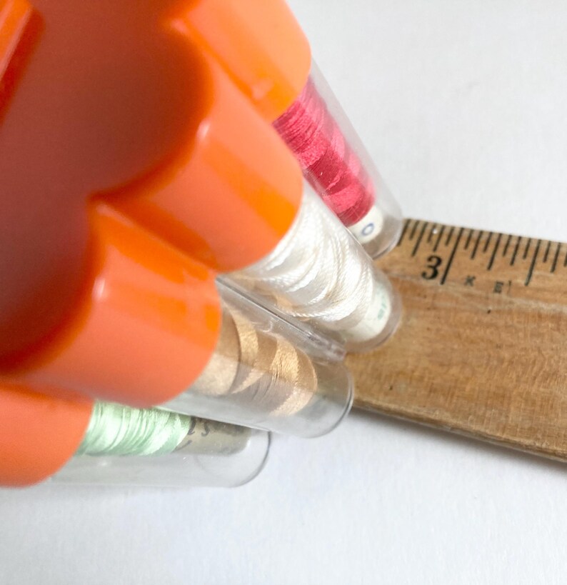 May include: An orange sewing kit with several clear tubes containing spools of thread in various colors, including red, white, gold, and green. A wooden ruler is positioned next to the tubes, showing the 3-inch mark.
