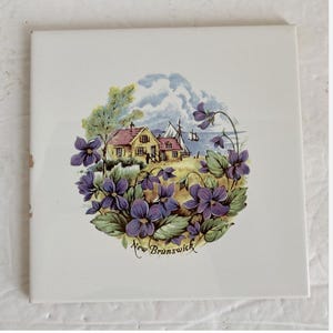 May include: Square ceramic tile with a scenic illustration of a coastal village surrounded by purple flowers. The image includes houses, sailboats, and the text "New Brunswick".