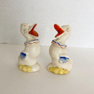 May include: A pair of vintage ceramic duck salt and pepper shakers. The ducks are white with orange beaks, blue and yellow accents, and a red, white, and blue collar. The salt shaker has holes on top.