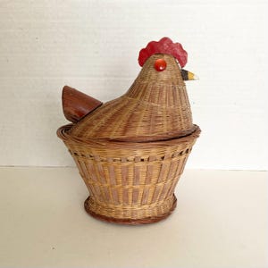 May include: A woven wicker basket shaped like a chicken with a removable lid. The basket features a natural tan color, a red painted wooden comb, a small orange bead for an eye, a pointed beak, and a dark brown tail piece attached to the lid.
